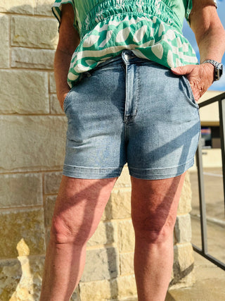 A person wearing mid-rise denim trouser shorts and a green top standing in front of a stone wall.