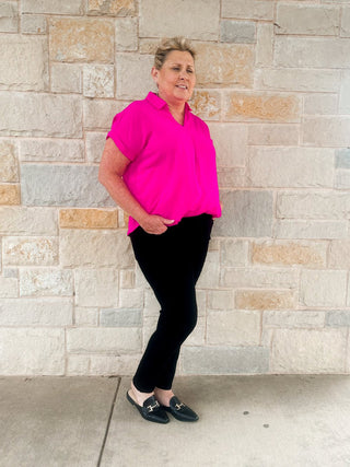 A person standing against a stone wall, wearing a pink mid-rise slim fit jean shirt and black pants, with their hands in their pockets.