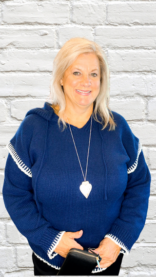 Woman wearing a blue sweater with white trim against a light gray brick wall.