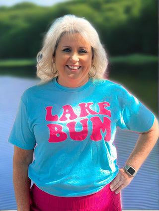 Woman wearing a blue t-shirt with 'LAKE BUM' text standing by a lake.