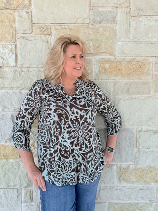 Woman wearing a patterned blouse and jeans standing against a stone wall.