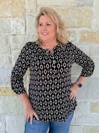 Woman wearing a black and white patterned blouse against a stone wall.