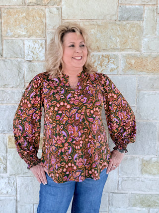 Woman wearing a patterned blouse and jeans standing against a stone wall.