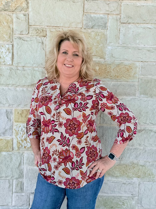 Woman wearing a floral blouse and jeans standing against a stone wall.