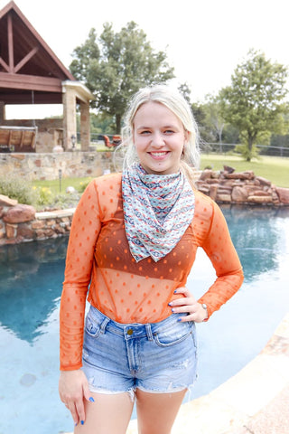 A woman wearing a long sleeve bronze mesh top and blue denim shorts, standing by a poolside.