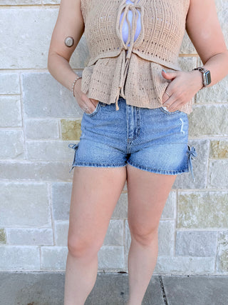 Person wearing a beige woven vest and denim shorts against a stone wall.
