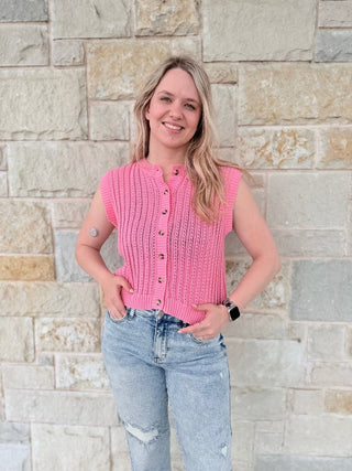 Woman wearing a pink sleeveless knitted top and light blue jeans standing against a stone wall.