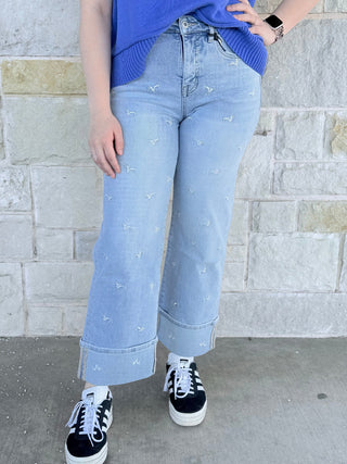 Person wearing light blue jeans with small white patterns, a blue top, and black and white sneakers against a stone wall.