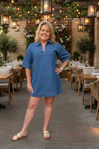 Woman in a blue dress standing in an outdoor dining area with decorative lights and plants.