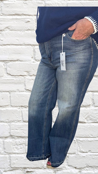 A pair of dark blue high-rise wide straight leg jeans with a tag on the back, displayed against a white brick wall background.