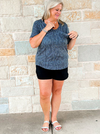 A woman wearing a black ribbed top with a cream boot stitch design, standing in front of a stone wall. She is also wearing black shorts and white sandals.