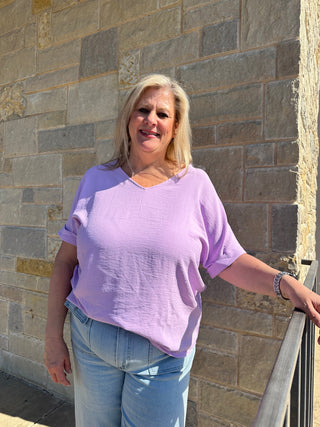 A woman wearing a purple v-neck blouse with dolman sleeves, standing in front of a stone wall.