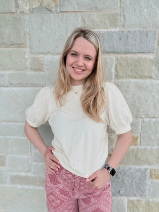 Woman wearing a white blouse and pink patterned pants standing against a stone wall.