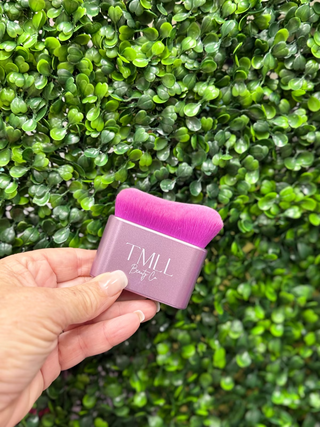 Hand holding a pink and purple beauty tool with 'TMLL' branding against a green leafy background