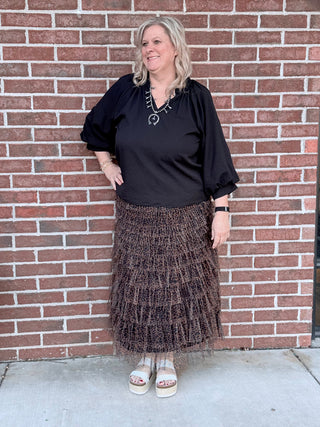 Woman wearing a black top and patterned skirt standing against a brick wall.