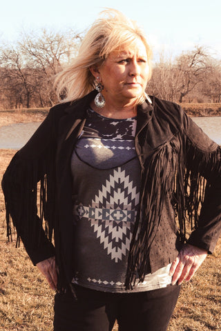 A person posing outdoors wearing a black velvet jacket with long fringe and decorative rhinestone embellishments, paired with a gray top with a geometric pattern and black pants.