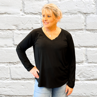 A woman wearing a black long sleeve top with a round neckline, standing against a brick wall backdrop.