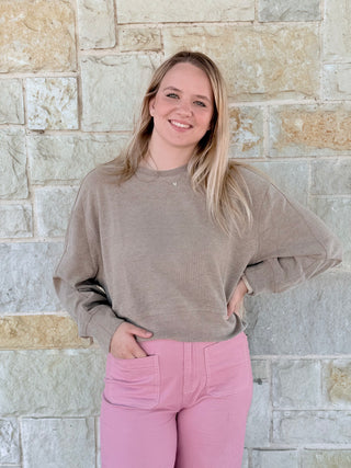 Woman wearing a beige sweater and pink pants standing against a stone wall.