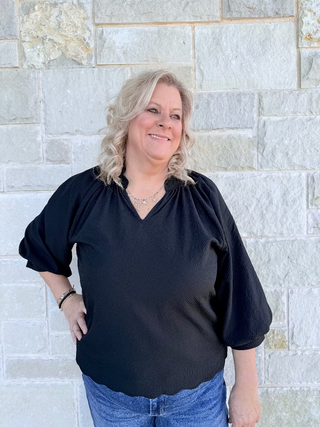 Woman wearing a black blouse and blue jeans standing against a stone wall.