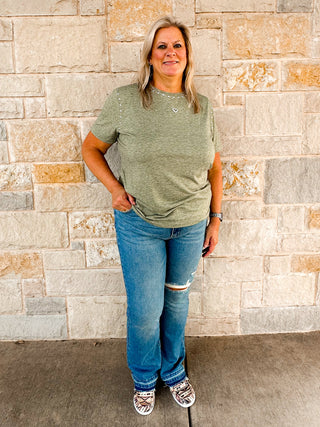 A woman standing against a stone wall, wearing a green t-shirt with studs around the collar and sleeves, paired with blue jeans.