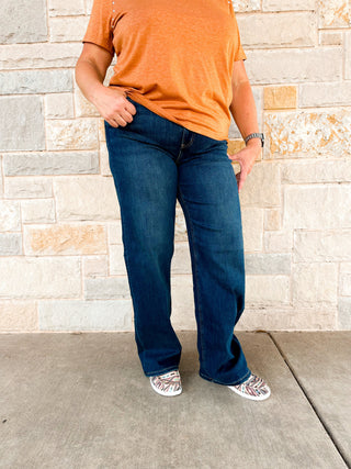 A person standing against a stone wall wearing high-waisted straight-leg jeans and an orange top.