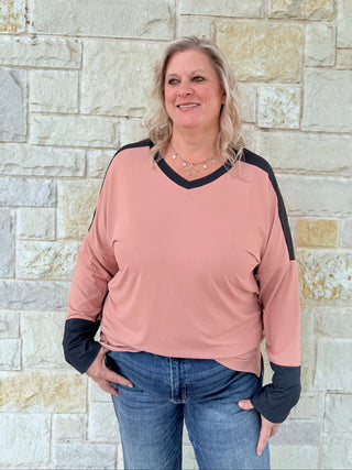 Woman wearing a pink long-sleeve top with black sleeves against a stone wall.