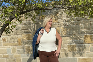 Woman wearing a white sleeveless top and brown pants against a stone wall.