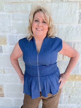Woman wearing a blue denim vest over a patterned dress against a light stone wall.
