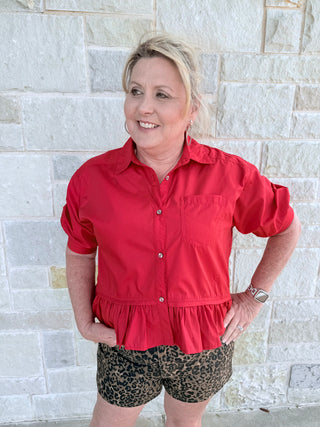 Woman wearing a red shirt and leopard print shorts standing against a light stone wall.