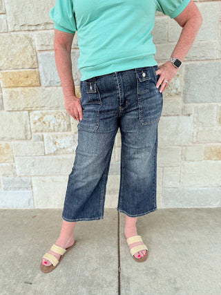 Person wearing a turquoise shirt and blue jeans standing against a stone wall.