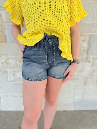 Person wearing a yellow crochet top and denim shorts against a stone wall background
