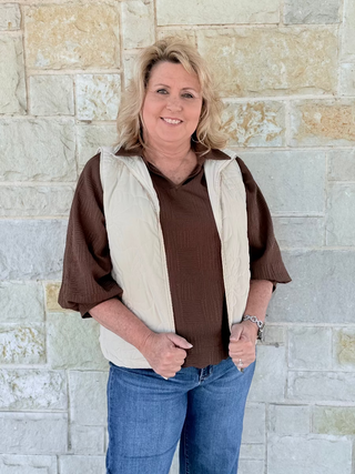 Woman wearing a brown shirt and beige vest standing against a stone wall.