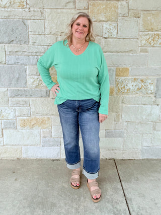 Woman wearing a green sweater and blue jeans standing against a stone wall.