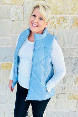A person wearing a blue vegan leather puffer vest with a round neckline and sleeveless design, paired with a white top and black pants.