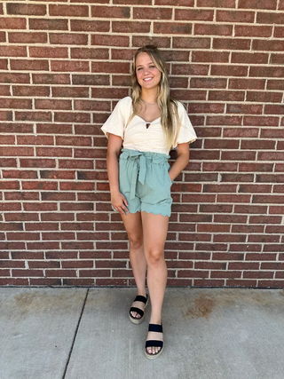 Woman standing against a brick wall wearing a white top and light green shorts.