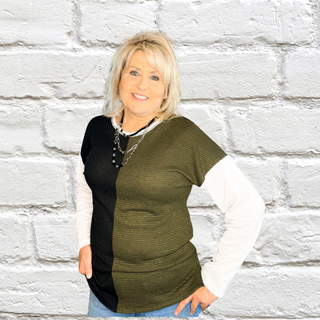A woman standing against a white brick wall background, wearing a waffle knit color block sweater with one half in black and the other half in hunter green, paired with a white top and blue jeans.