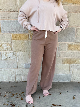 Person wearing a beige hoodie and brown sweatpants standing against a stone wall.