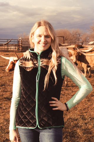A woman standing in a field with cows in the background, wearing a black velvet quilted vest with turquoise lining and a zipper.