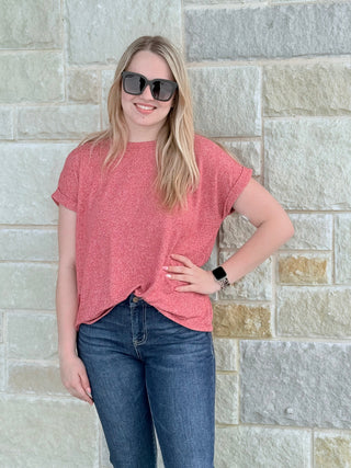 Woman wearing a red t-shirt and blue jeans standing against a stone wall.