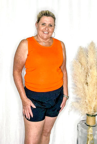 A smiling woman standing, wearing an orange tank top and black shorts, with a feather decoration in the background.