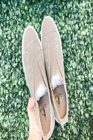 A pair of beige clog-style slippers with a textured sole, held against a green fuzzy background.