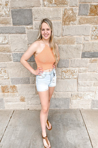 A knitted halter crop top in orange, displayed flat with a pair of denim shorts and a straw hat in the same color palette.