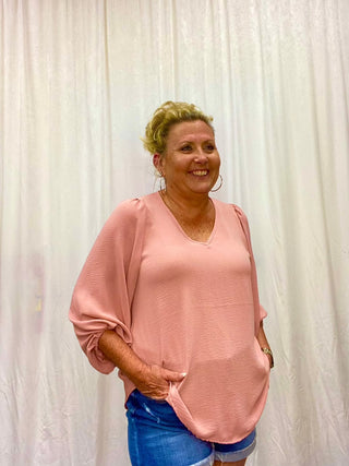 A woman standing with one hand on her hip, wearing a pink V-neck cinched sleeve top and blue denim shorts.